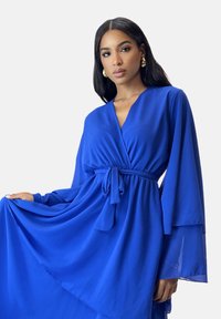 Lang blå chiffon kjole med V-hals, lange klokkeærmer og bindebånd i taljen. Stoffet er flydende med en blød tekstur og let glans.