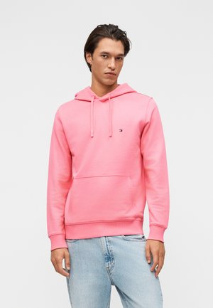 Mies, jolla on päällään vaaleanpunainen hupullinen collegepaita edessä taskulla ja vaaleansiniset farkut, seisoo yksinkertaista vaaleaa taustaa vasten.