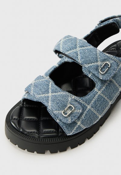 Denim schuifsandaal met een gequilt zwarte voetbed, verstelbare bandjes en zilveren hardware accenten. Blauw denim met een wit ruitpatroon.