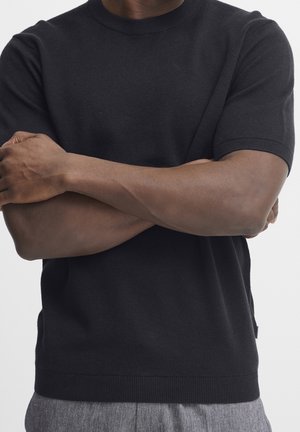 Homme portant une chemise noire à manches courtes, les bras croisés sur la poitrine, debout devant un fond clair uni.