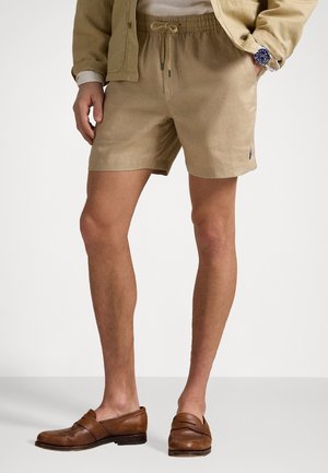 Beżowe lniane szorty z elastycznym pasem i sznurkiem do ściągania. Posiadają boczne kieszenie oraz małe logo na brzegu. Noszone z brązowymi mokasynami.