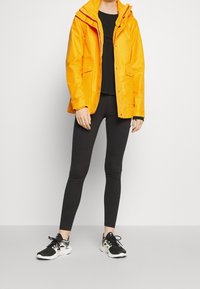 Giacca impermeabile gialla con cappuccio, chiusure a zip e a pressione, indossata sopra una maglietta nera. Leggings neri e scarpe da corsa completano l'outfit.