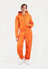 Oranje zip-up hoodie met voorzakken en bijpassende joggingbroek. Zachte stof met elastische manchetten. Logodetail linksboven. Witte sneakers.