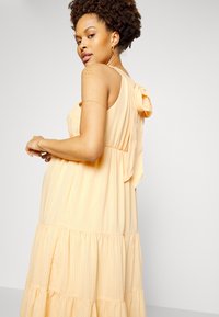 Vestuário maxi em amarelo claro, com camadas em tecido texturizado e detalhe de laço nas costas, apresentando um corte descontraído e alças ajustáveis.