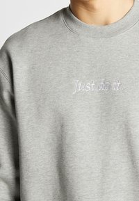 Šedá mikina s kulatým výstřihem, se vyšitým textem "Just do it." ve světle fialové barvě. Materiál má měkký, texturovaný vzhled.