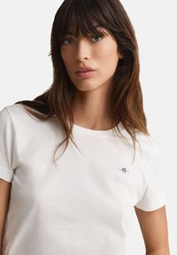 White cotton t-shirt with a crew neckline and short sleeves, featuring a small, embroidered logo on the left chest. Smooth texture.