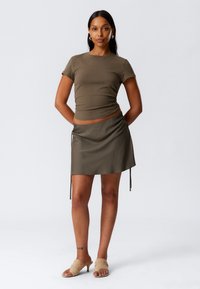 Femme se tenant avec les mains derrière le dos, portant un haut à manches courtes olive, une jupe assortie avec des liens sur les côtés, et des sandales à talons beiges.