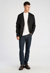 Czarny prążkowany kardigan, zapinany na guziki, noszony na białej koszulce. Dopełnieniem stroju są niebieskie dżinsy i brązowe buty. Prosty, nowoczesny design.