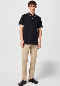 Černé polo tričko s bílým lemem, krátké rukávy a tříknoflíkovou légou. Nosit s béžovými kalhotami a černými slip-on botami.