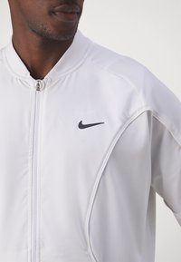 Weiße Nike-Jacke mit Reißverschluss vorne, geripptem Kragen und schwarzem Swoosh-Logo auf der linken Brust. Glatte Textur und taillierte Passform.