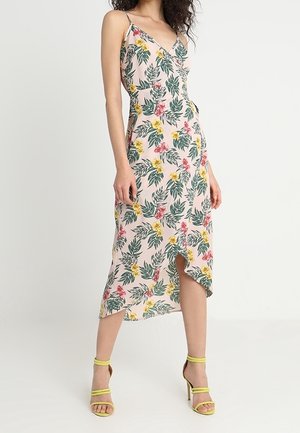 Vrouw draagt een mouwloze, roze, bloemen midi-jurk met gele en roze bloemen en groene bladeren, gecombineerd met gele sandalen met hoge hakken.