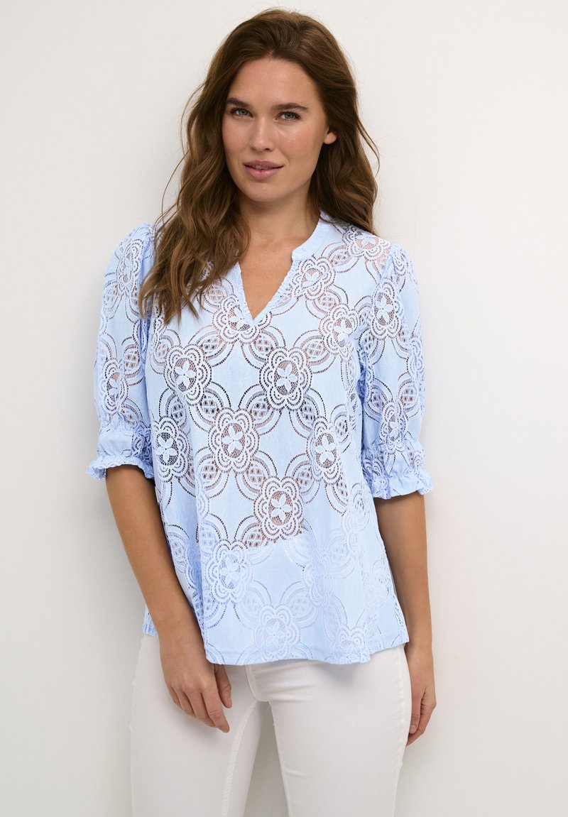 Blusa de encaje azul claro con patrones florales, escote en V y mangas abullonadas, hecha de un tejido delicado y texturizado.