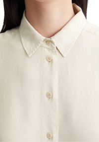Camicia beige con bottoni, dotata di un colletto classico, presenta sei piccoli bottoni coordinati e una texture liscia e tessuta.