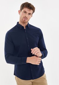 Marineblauwe button-up shirt met een slim fit, lange mouwen en subtiel logo op de borst. Gladde stof met afgeronde zoom en knopenmanchetten.