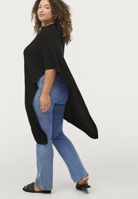 Sort poncho-lignende top med vidde ærmer, parret med lyseblå flared jeans. Toppen har en løs pasform og en struktureret kantdetalje.