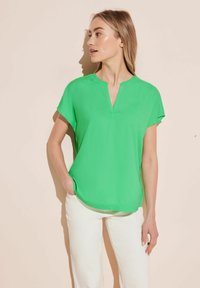 Blusa verde a maniche corte con scollatura a V, realizzata in tessuto morbido. Abbinata a pantaloni bianchi, con una vestibilità morbida e una leggera curvatura all'orlo.