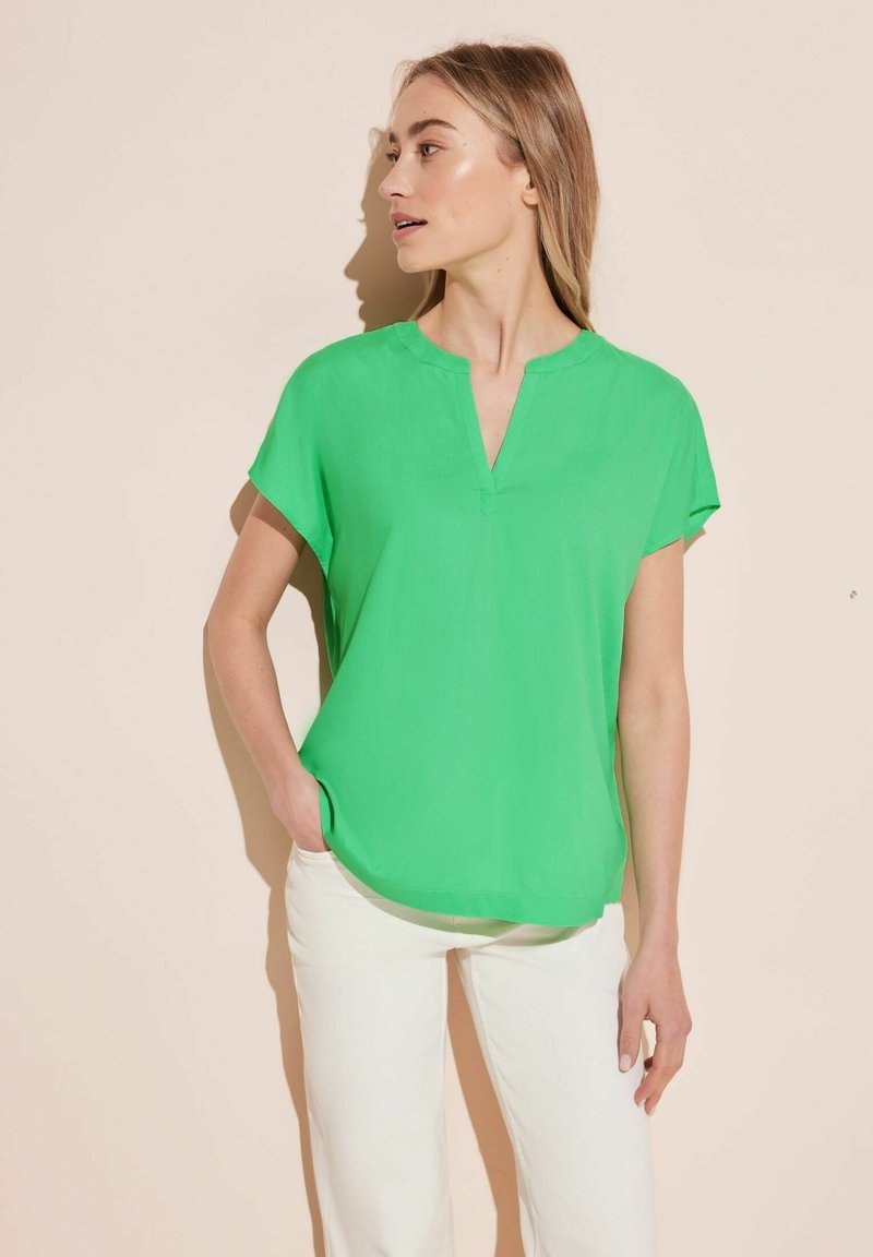 Blusa verde a maniche corte con scollatura a V, realizzata in tessuto morbido. Abbinata a pantaloni bianchi, con una vestibilità morbida e una leggera curvatura all'orlo.