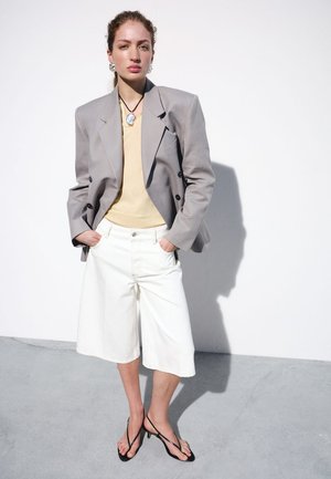 Mujer con blazer gris claro de gran tamaño, top beige, shorts blancos hasta la rodilla, tacones negros con tiras, de pie contra una pared blanca con las manos en los bolsillos.