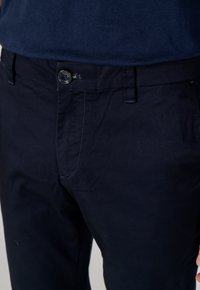 Pantaloni di cotone blu navy con chiusura a bottone, frontale piatto e tasche laterali. Presentano una sottile texture e cuciture a contrasto.