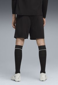 Sorte atletiske shorts parret med sorte knæhøje sokker med en hvid stribe. Personen bærer hvide fodboldstøvler med et logo.