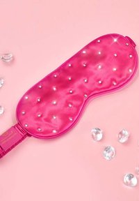 Mascherina da notte in raso rosa con strass sparsi, forma sagomata e fascia elastica. Circondata da accenti trasparenti simili a cristalli su uno sfondo rosa.