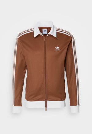 Chaqueta deportiva marrón con cuello y puños blancos, cierre de cremallera y tres rayas blancas a lo largo de las mangas. Logo de Adidas en el pecho.