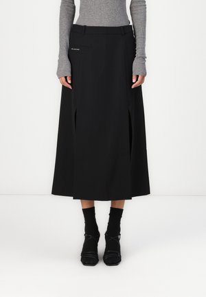 Svart midi skjørt med sidelommer, beltehemper og frontlomme. Laget av glatt stoff med en subtil glans. Kombinert med svarte ankelstrømper og sko.