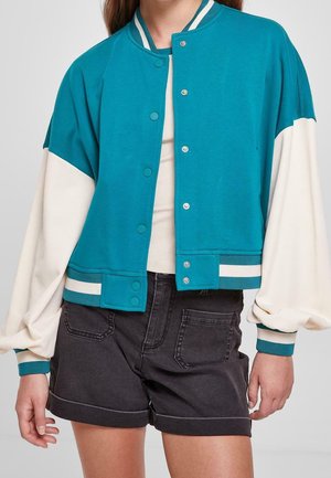 Teal og creme varsityjakke med trykknapper, kombineret med sorte shorts med opsmøg og en beige top, båret af en stående person.