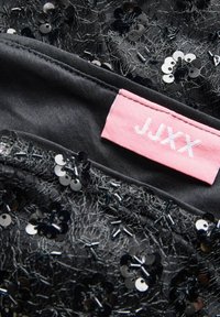 Melnā auduma ar sekmēm un pērlītēm. Uz etiķetes redzams "JJXX" baltā krāsā uz rozā fona. Tekstūrveida, slāņainais dizains ar ziedu formām.