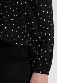 Black long-sleeve blouse with silver polka dots and elastic cuffs, worn tucked into black pants, showing a small decorative pin near the waist.