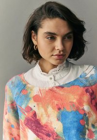 Sveter z cashmere-umeleckou zmesou má farebný abstraktný vzor s odtieňmi ružovej, modrej a oranžovej. Je vrstvený cez bielu košeľu s krajkovým lemom.