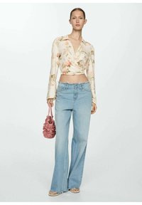Žena v kvetinovom zaviazanom crop top, svetlomodrých vysoko vyvýšených širokých džínsoch, béžových šľapkách a drží ružovú textúrovanú kabelku.