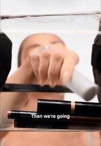 Une main atteignant un récipient transparent contenant des produits de maquillage noirs et beiges, le texte superposé indique "Then we're going".