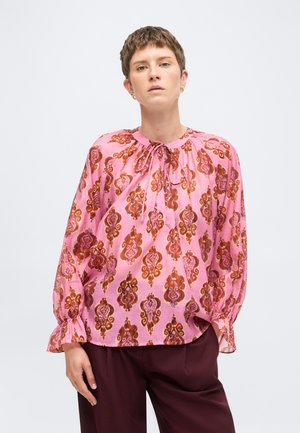 Femme aux cheveux courts portant un chemisier rose à motif avec un col à nouer et des manches longues, associé à un pantalon bordeaux foncé.