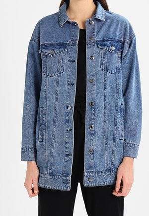 Denim jacket with button front, chest flap pockets, and side pockets worn over a black top and black pants.