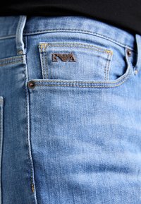 Lyseblå denimjeans med bronsebeslag, med et brodert logo på baklommen og synlige sømdetaljer.