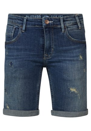 Barn står med hænderne i lommerne iført en marineblå T-shirt med grafik, opkørte denimshorts og lyse slip-on sko.