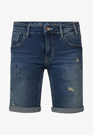 Barn står med hænderne i lommerne iført en marineblå T-shirt med grafik, opkørte denimshorts og lyse slip-on sko.