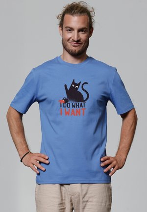 Blauwe katoenen T-shirt met een afbeelding van een zwarte kat en de tekst "IK DOE WAT IK WIL" in vette rode letters. Korte mouwen, ronde hals.