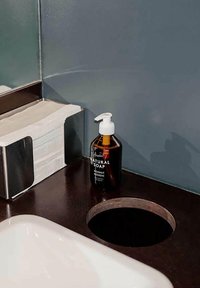 Piano di lavoro scuro con un'apertura rotonda, lavandino bianco, dispenser di sapone marrone con l'etichetta "Sapone Naturale" e un portatavoliere in argento.