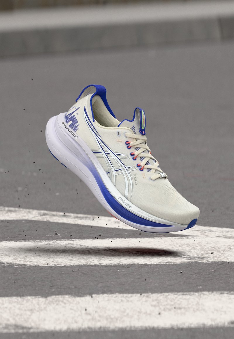 Pantof de alergare alb și albastru în aer deasupra unui drum de asfalt gri cu dungi albe vopsite, prezentând detalii ale părții laterale și ale tălpii.