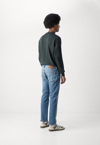 Donkergroene sweatshirt, blauwe jeans met leren patch en groen gestreepte sneakers; casual ontwerp met een relaxte pasvorm en lichte omslag bij de enkels.