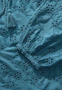 Blusa teal con ricami floreali a rilievo. Le maniche hanno un polsino elastico e un dettaglio con nappine vicino al collo.