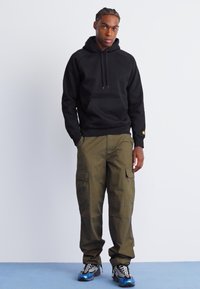 Sweatshirt à capuche noire, pantalon cargo vert olive avec poches latérales, associé à des baskets avec des accents bleus et argentés.