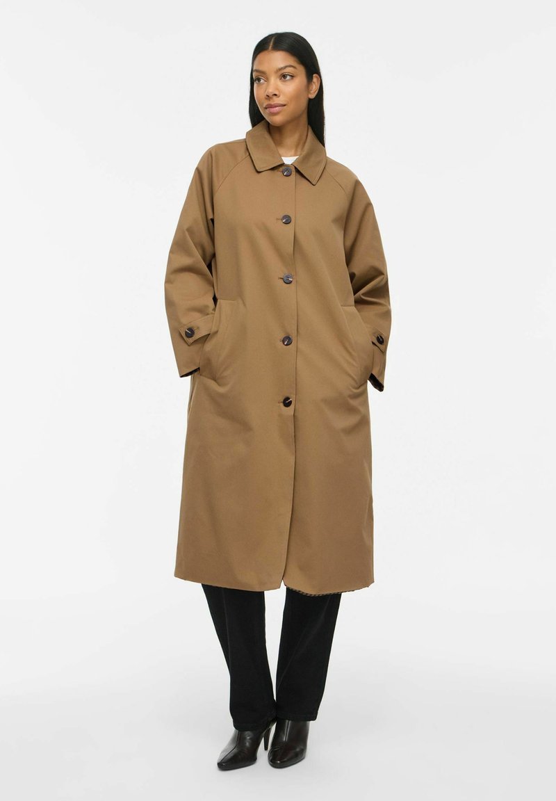 Femme portant un long manteau marron boutonné avec col, mains dans les poches, pantalon noir et bottes à talons noires sur un fond uni.