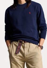 Mörkblå sweatshirt med rund halsringning och röd logga. Tillsammans med beige byxor som har ett randigt bälte och synliga fickdetaljer.