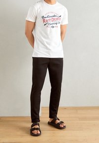 Męska biała koszulka bawełniana z czerwonym i niebieskim graficznym tekstem. Czarne spodnie skinny o gładkiej teksturze oraz płaskie sandały z regulowanymi paskami.