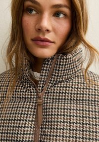Bruine en zwarte houndstooth-gepatroneerde jas met een hoge kraag, voorzien van een voorknof en een gestructureerde stof.