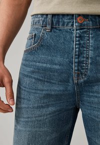 Jeans de mezclilla azul con un corte recto, costuras naranjas, un bolsillo frontal y un botón marrón en la cintura.