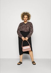 Femme aux cheveux bouclés portant un blouse à motifs floraux et une jupe noire, tenant un sac à main rose, se tenant devant un fond blanc uni.